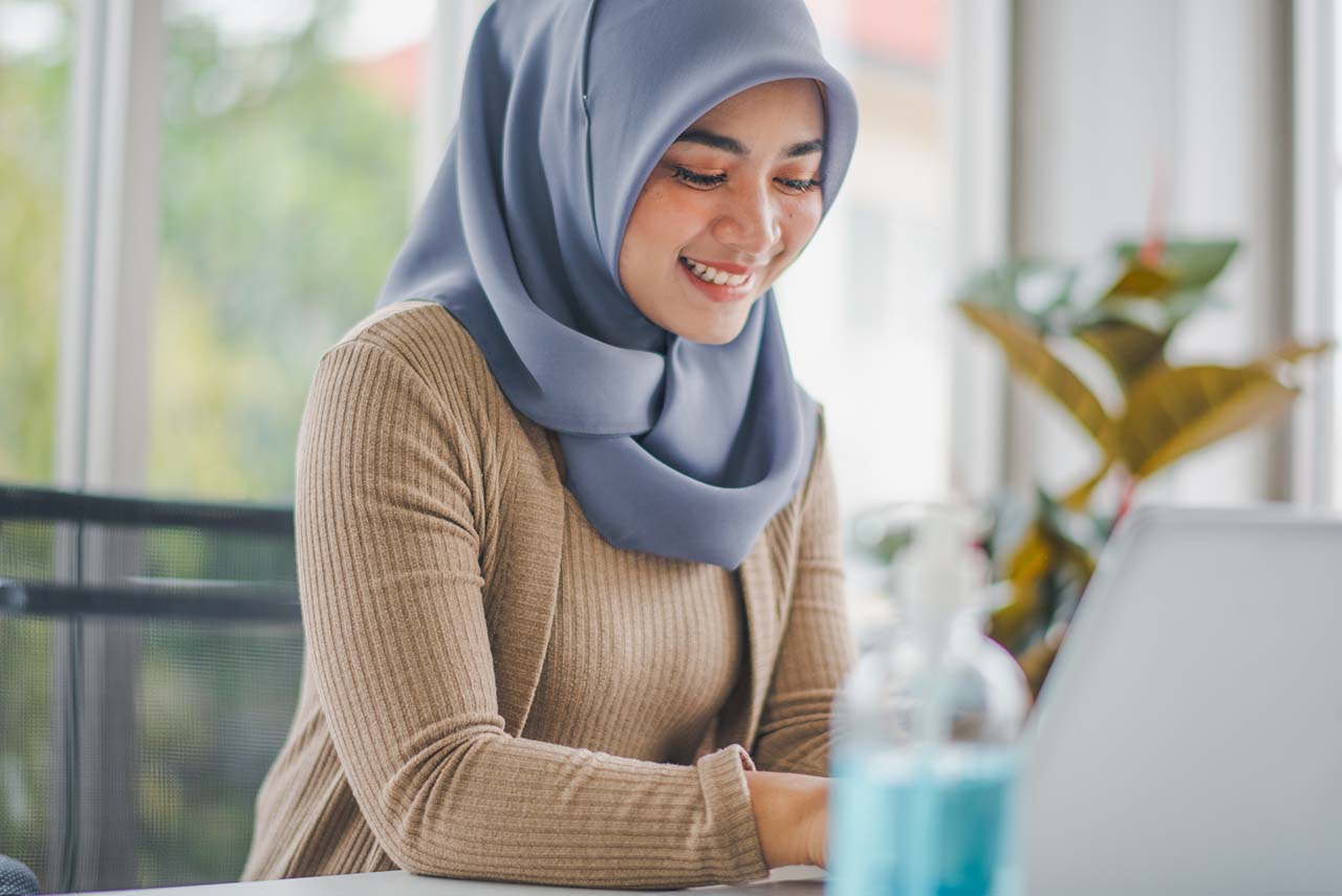 Young Islam Asian woman take hijab is washing hand by alcohol gel before use computer notebook to work in living room at home office which glad smiling and felling happy in COVID19 pandemic.